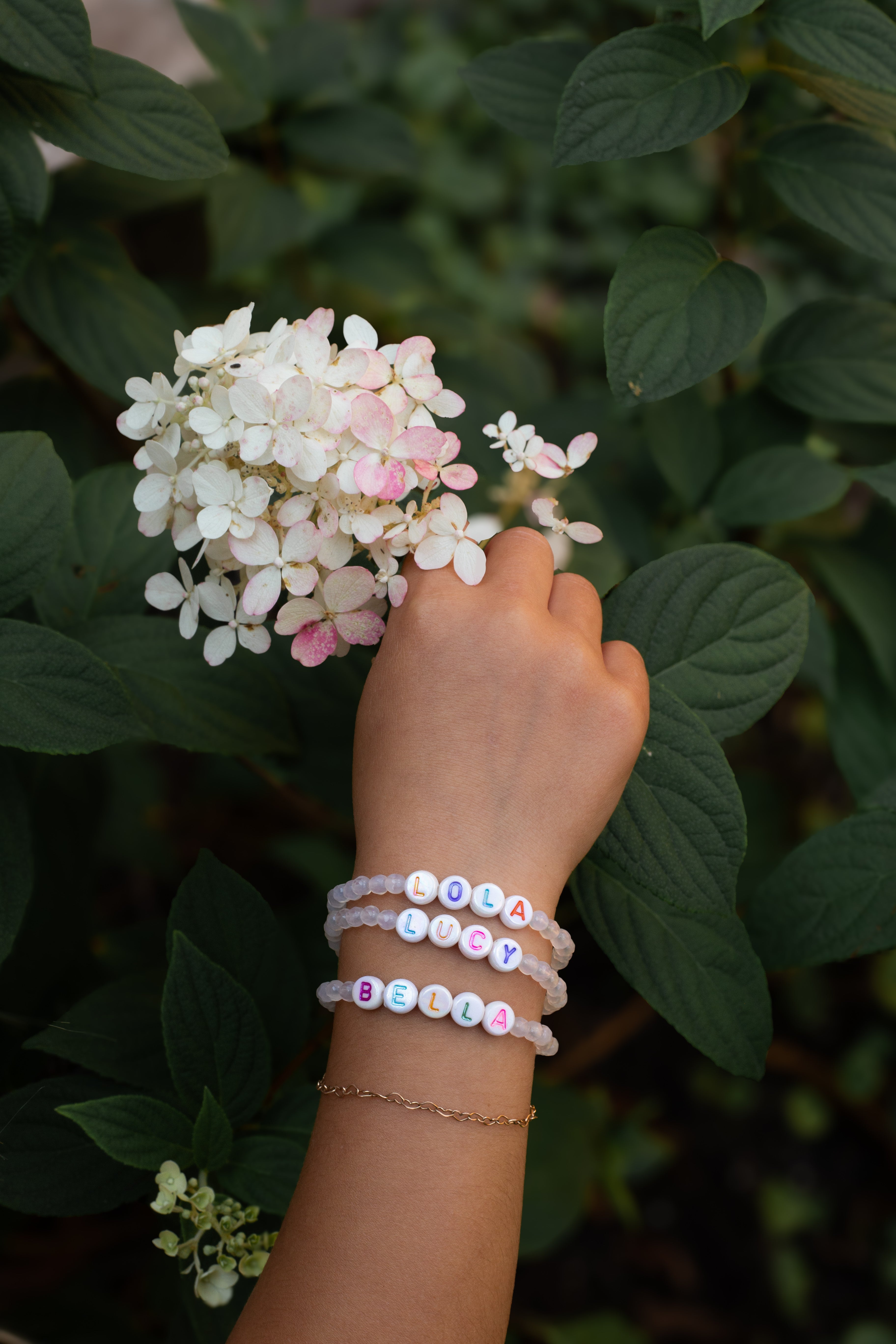 Hand wearing colorful beaded bracelets touching a cluster of white and pink flowers with green leaves.